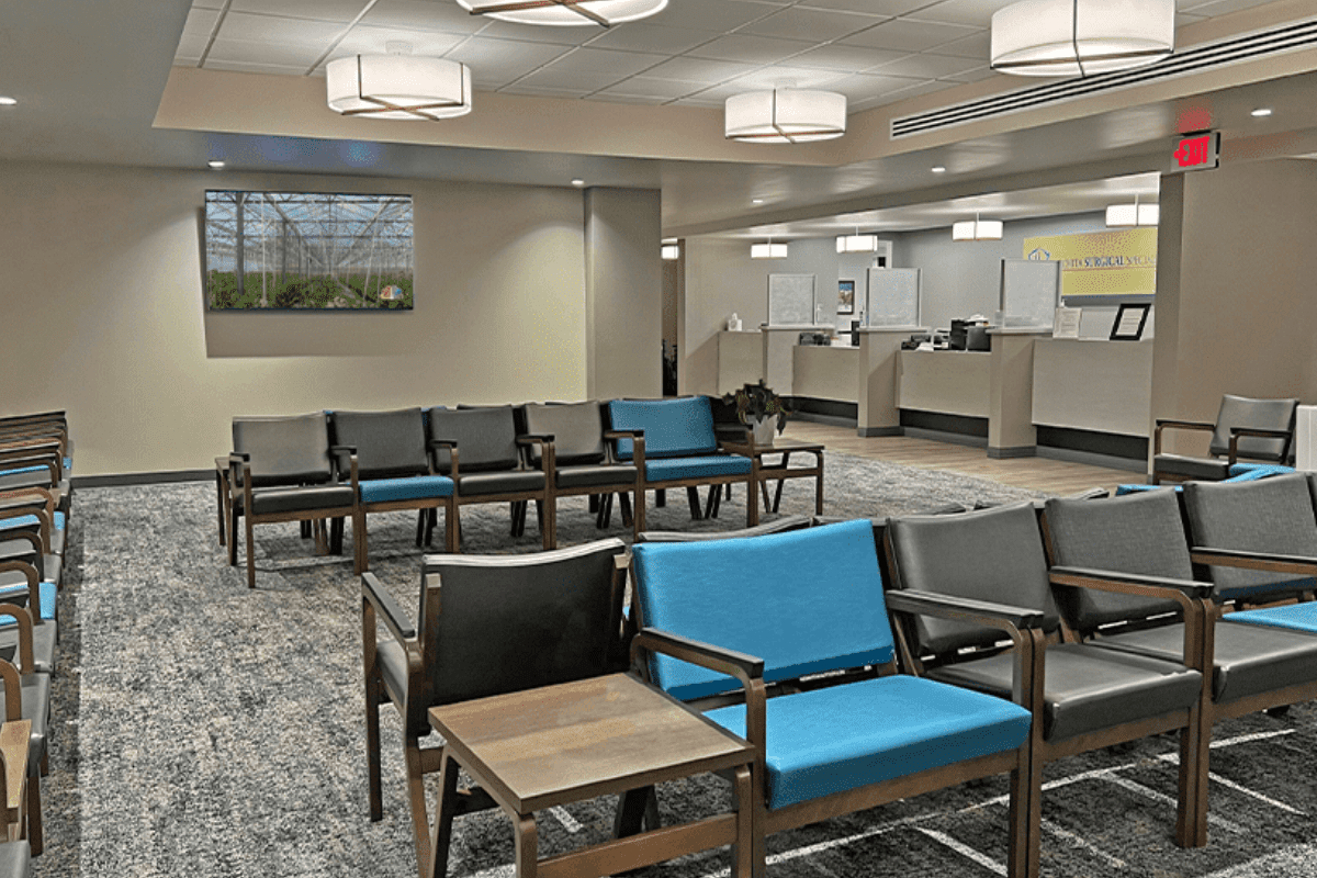 Waiting area with grey and blue chairs, a tv, and a reception desk in the back of the photo.