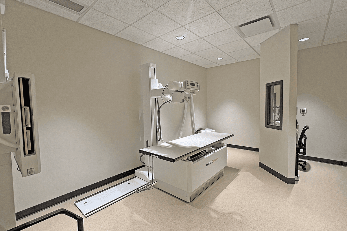 X-ray imaging room with full body scanning equipment and small workstation behind a lead-lined wall.