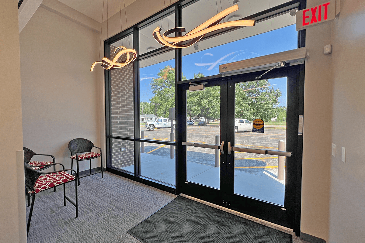 Interior view of a building with large windows overlooking a parking lot, featuring two black chairs with red patterned cushions and a unique, curved light fixture on the ceiling.