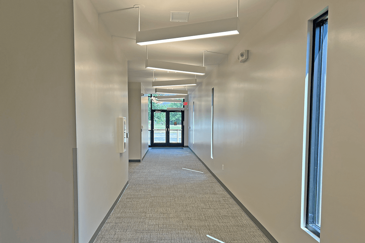 Long, narrow interior hallway with white walls, gray carpet, and modern, angled fluorescent lighting fixtures. A doorway is visible at the end of the hall.
