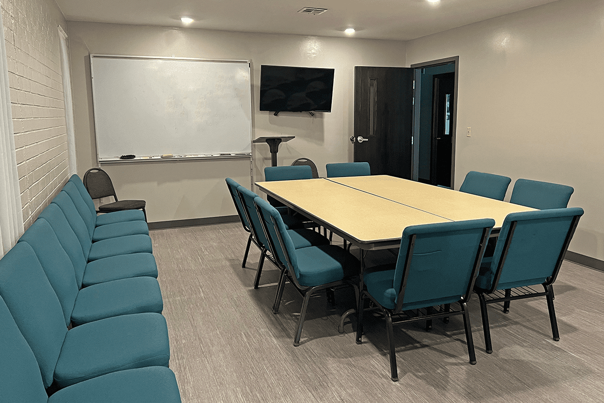 Meeting room with a rectangular conference table surrounded by teal chairs, a whiteboard, and a wall-mounted television.
