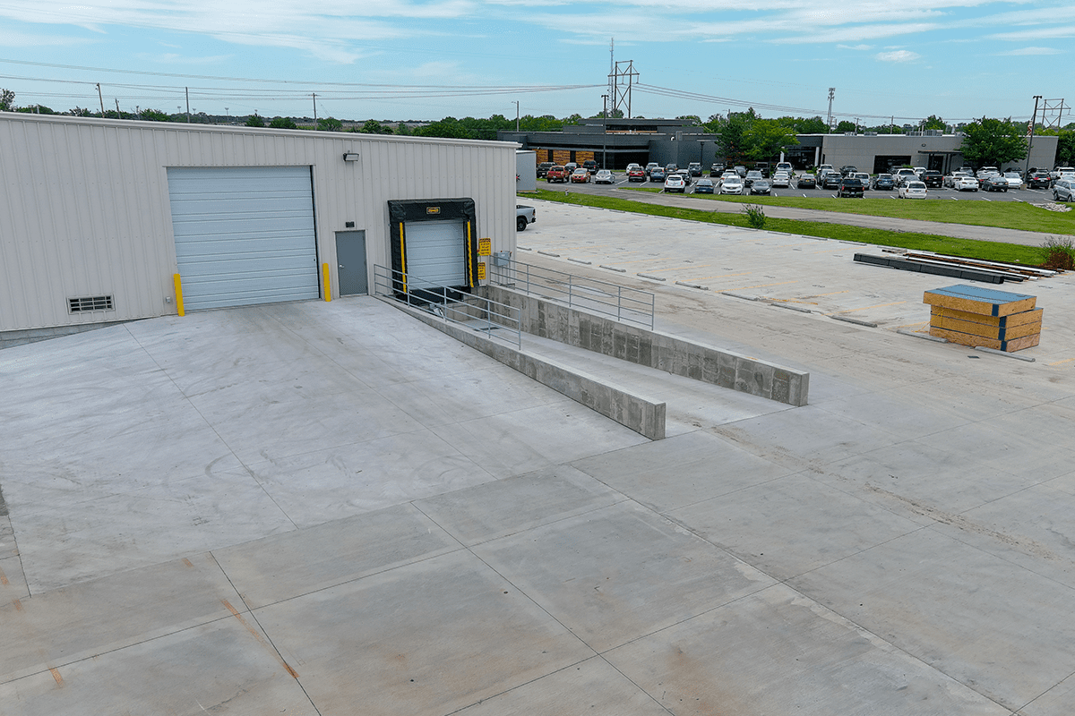 Commercial warehouse with a gray facade, multiple garage doors, and many parked vehicles in the background.